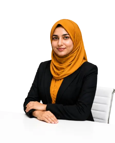A woman wearing a hijab is seated at a table, engaged in work with a laptop and a notepad beside her.
